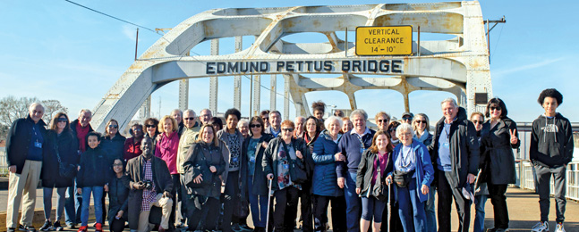 A long line of people stand in front of the Edmund Pettus Bridge
